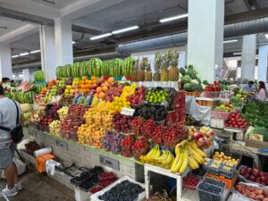 A vibrant market stall filled with a variety of fresh vegetables and fruits including watermelons, apples, bananas, and pineapples, with shoppers browsing the selection.